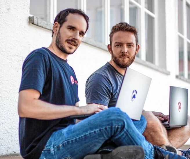 Die beiden Geschäftsführer von RedFox InfoSec sitzen mit aufgeklappten Laptops vor einem Gebäude.